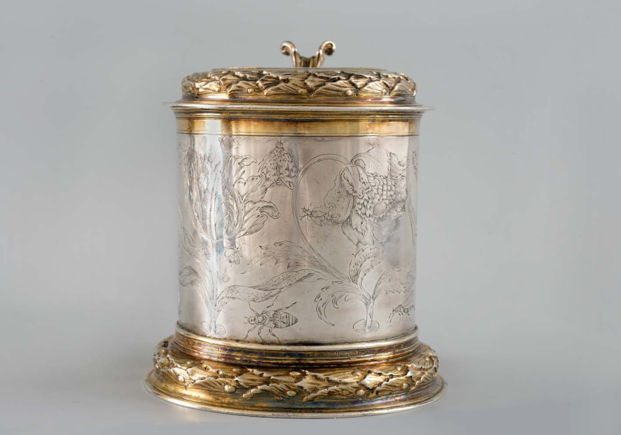 Srebrny kufel ze złotymi motywami zdobiony grawerunkiem. Na jego brzuścu znajdują się kwiaty oraz owady. 

