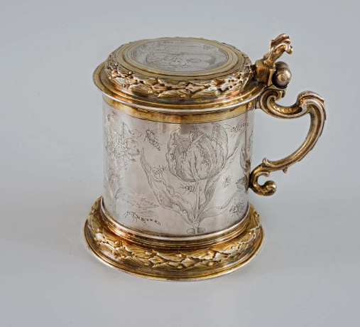 Srebrny kufel ze złotymi motywami zdobiony grawerunkiem. Na jego brzuścu znajdują się kwiaty oraz owady. 

