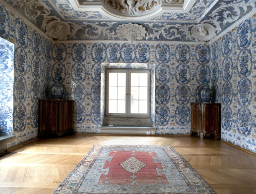 Pomieszczenie wyłożone biało-niebieskimi, bogato zdobionymi płytkami, na pierwszym planie znajduje się okno i niebiesko-czerwony dywan