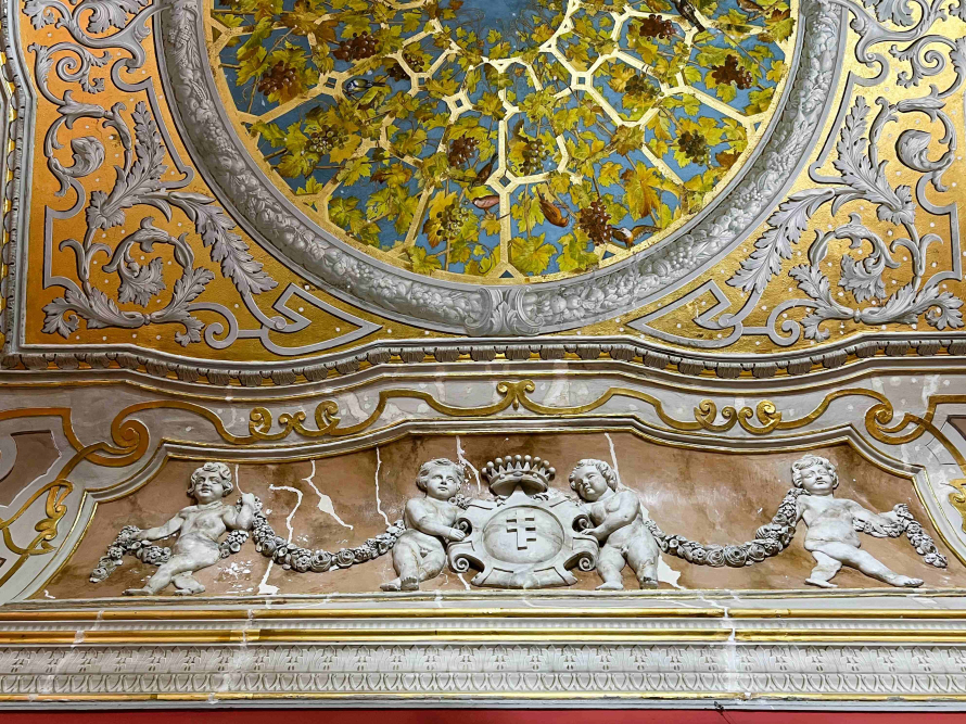 Fragment dekoracji rzeźbiarsko malarskiej z historycznego wnętrza pałacu. Widoczne płaskorzeźbione putta, herb Pilawa oraz malowany ążurowy otwór altany z wijąca się roślinnością.