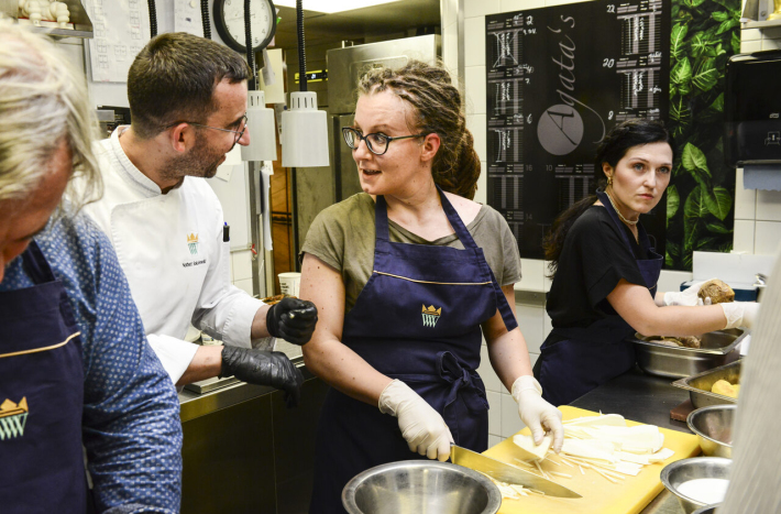 Czworo ludzi w kuchni w trakcie przygotowywania posiłku. Dwie osoby w centrum - mężczyzna i kobieta rozmawiają.