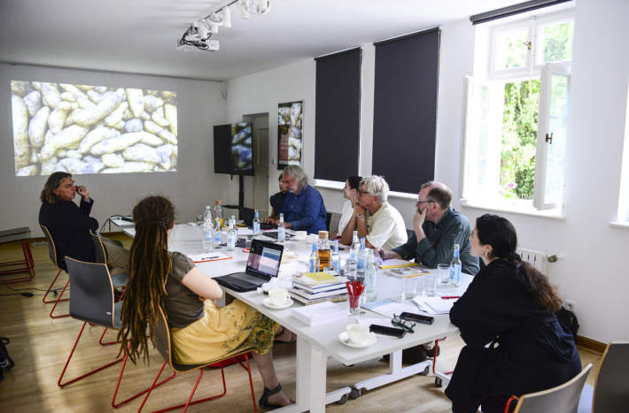 Grupa osób siedzi przy stole i jest w trakcie spotkania. Na ścianie widnieje prezentacja, o której opowiada jeden z uczestników.